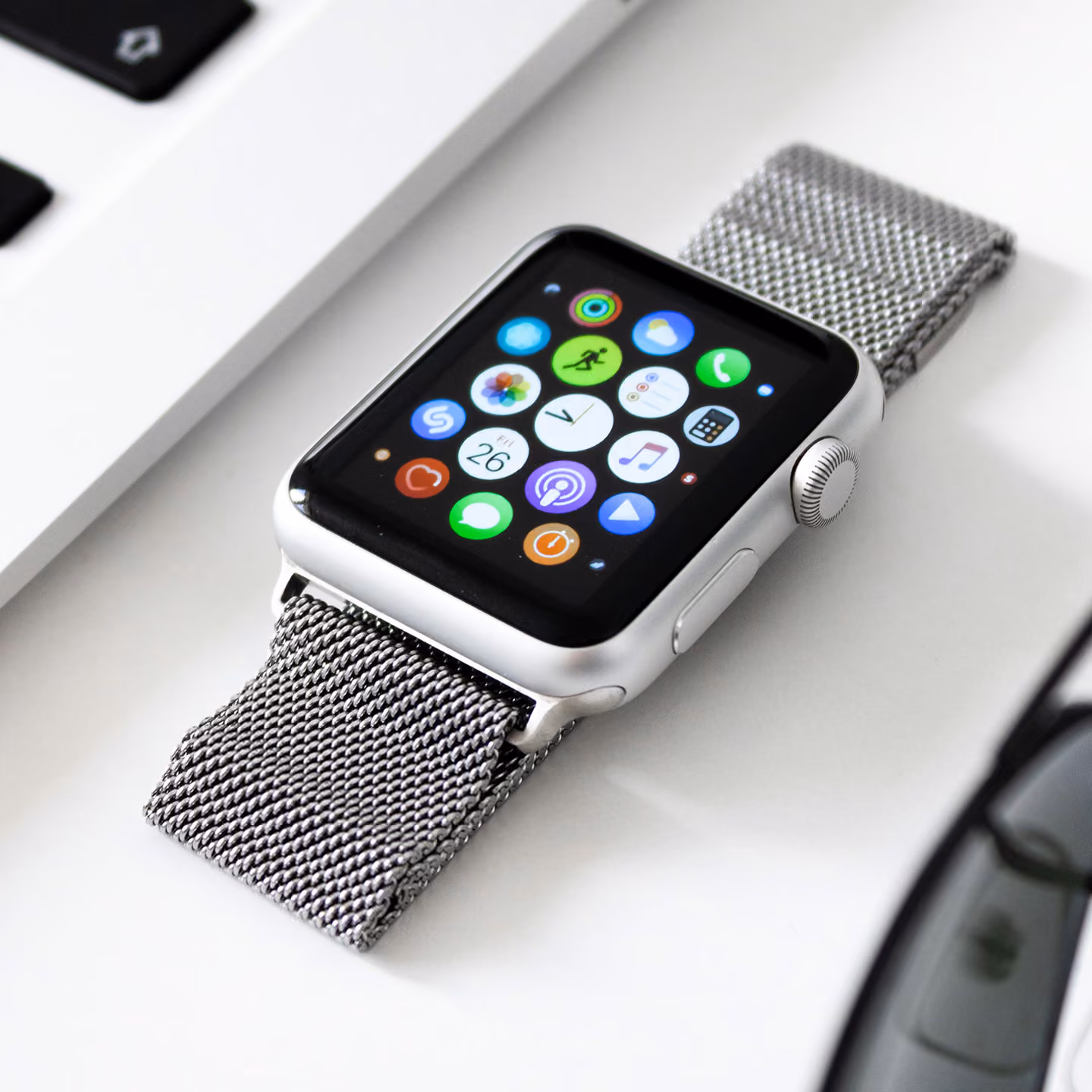 Silver smartwatch with a woven metal band showing app icons on its screen on a white surface.