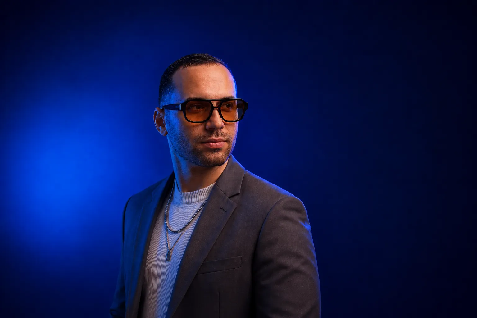 Man wearing orange-tinted glasses, a gray blazer, and layered necklaces on a blue background.